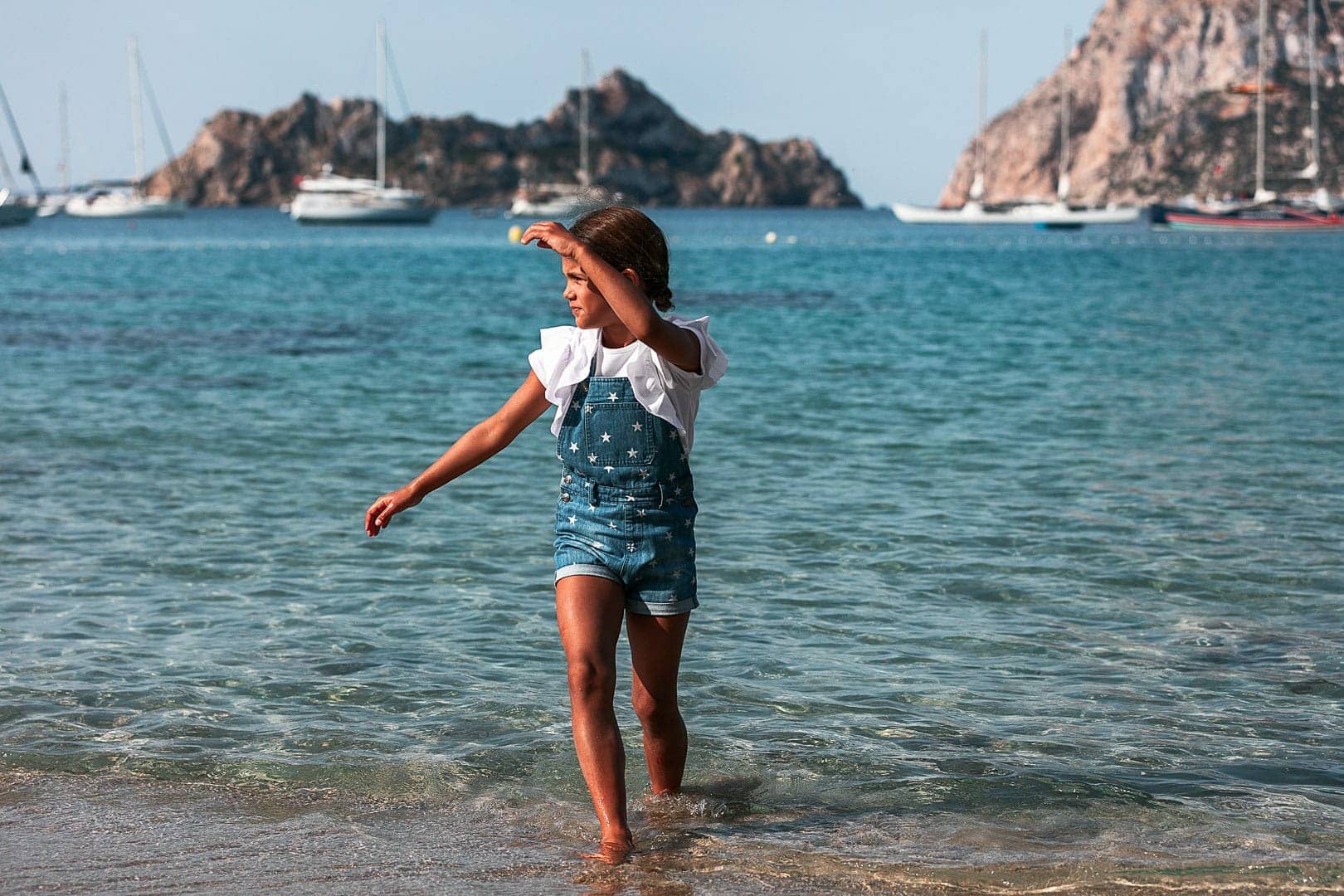 Photographic report of children's clothing with a girl walking on the shore of Cala Dort beach in Ibiza dressed in denim overalls
