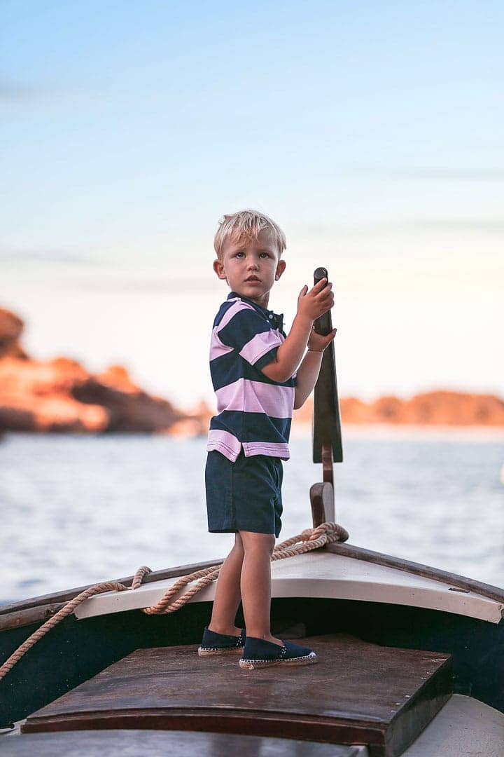 Retrato de un niño sonriente jugando sobre una barca vestido con camiseta rayada de color azul y pantalón azul en embarcadero de Ibiza