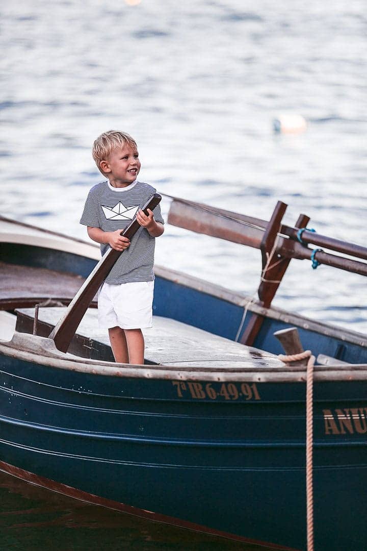 Retrato de un niño sonriente jugando sobre una barca vestido con camiseta rayada de color azul y pantalón blanco en embarcadero de Ibiza